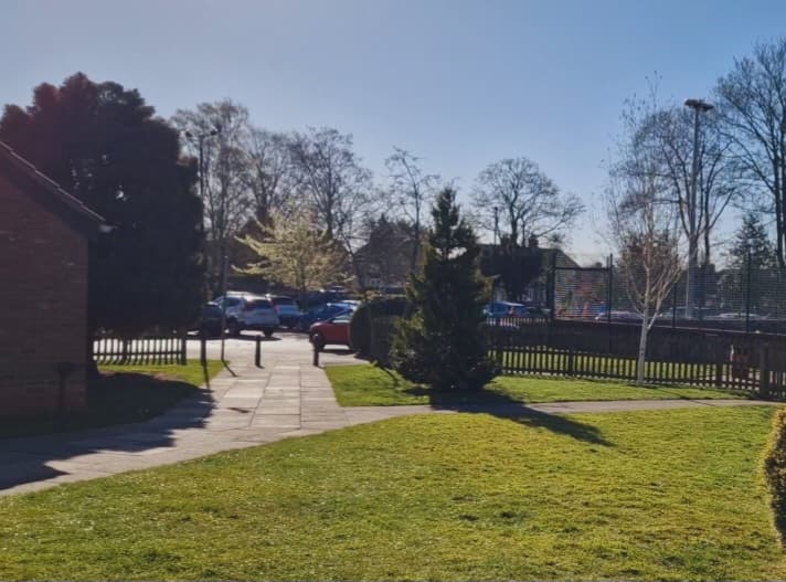 Sunny car park at Galtres Centre, surrounded by trees and grass, with parked cars and a clear blue sky.