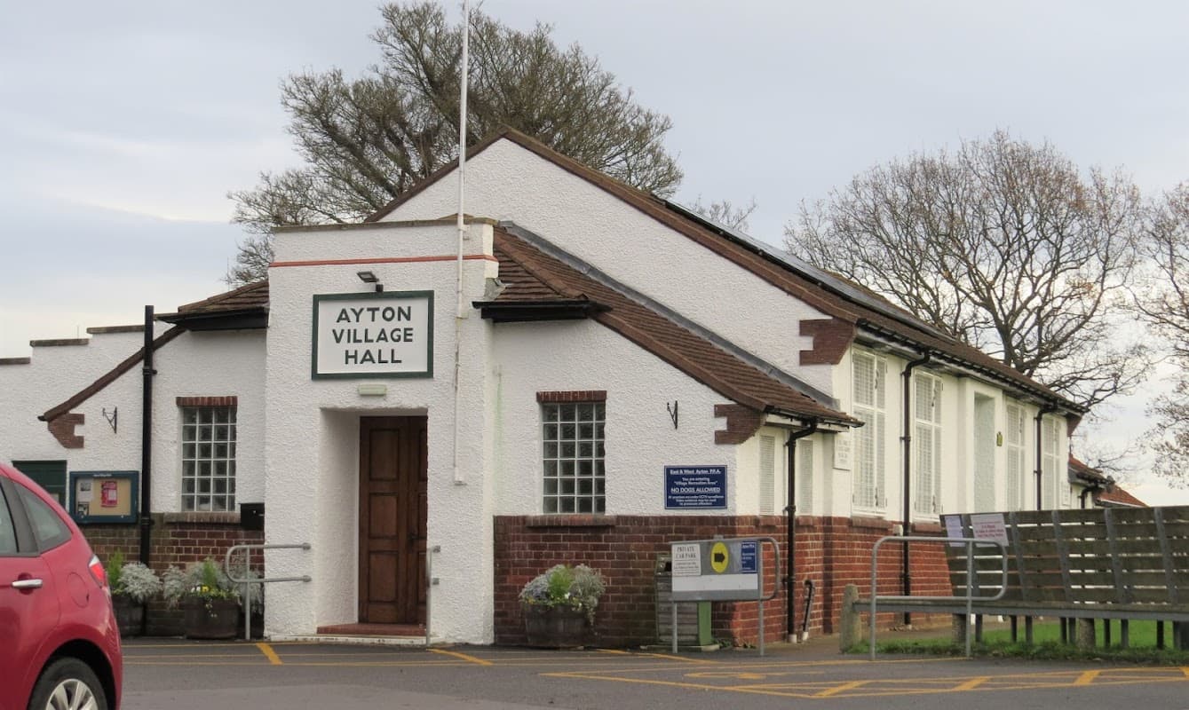 White building with a sign reading "Ayton Village Hall," surrounded by trees and a parking area.