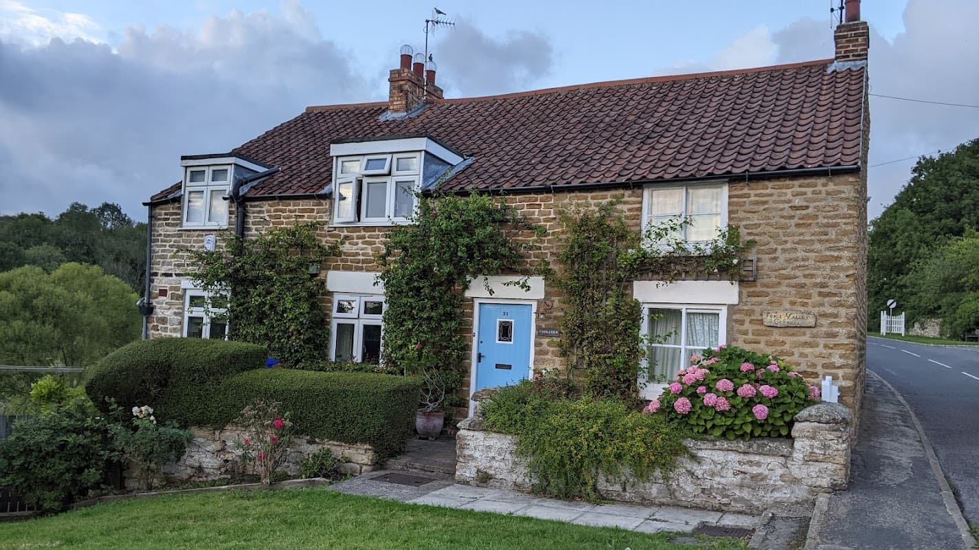 Charming stone cottage with a blue door, surrounded by greenery and flowers, set against a rural backdrop in East Ayton.