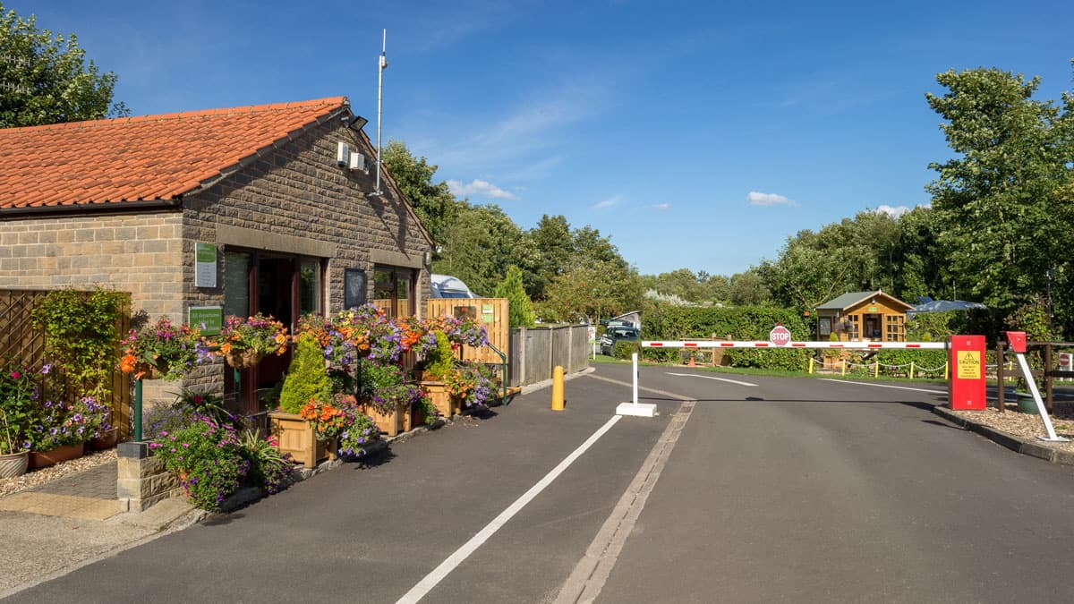 Campsite entrance with a stone building, flower pots, and a barrier, surrounded by greenery and blue sky.