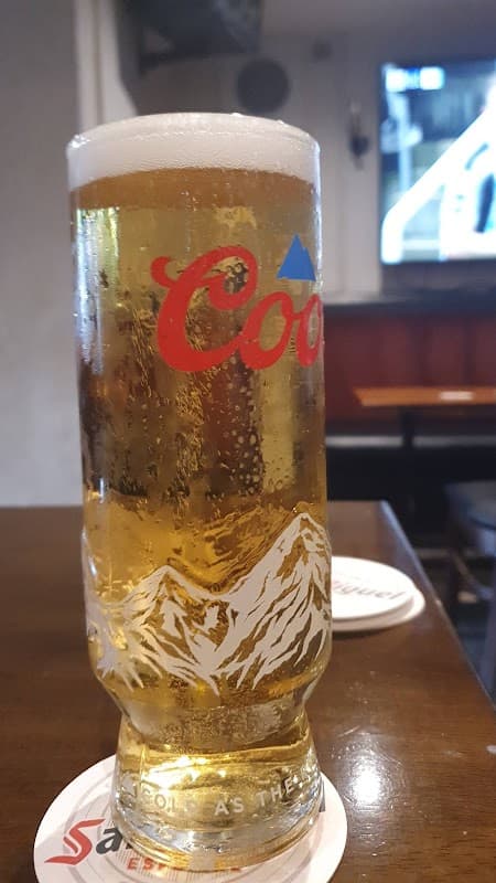 A tall glass of beer with a frothy top, sitting on a wooden table in a cozy pub setting.