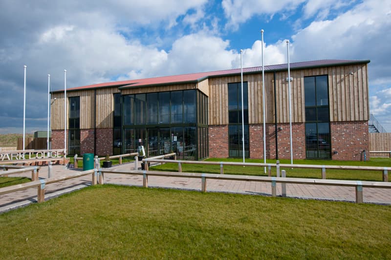 Modern building with wooden and brick facade, large glass windows, surrounded by green grass and flagpoles.