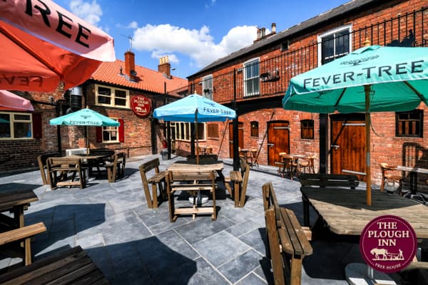 Outdoor seating area with wooden tables and colorful umbrellas at The Plough Inn, surrounded by brick buildings.