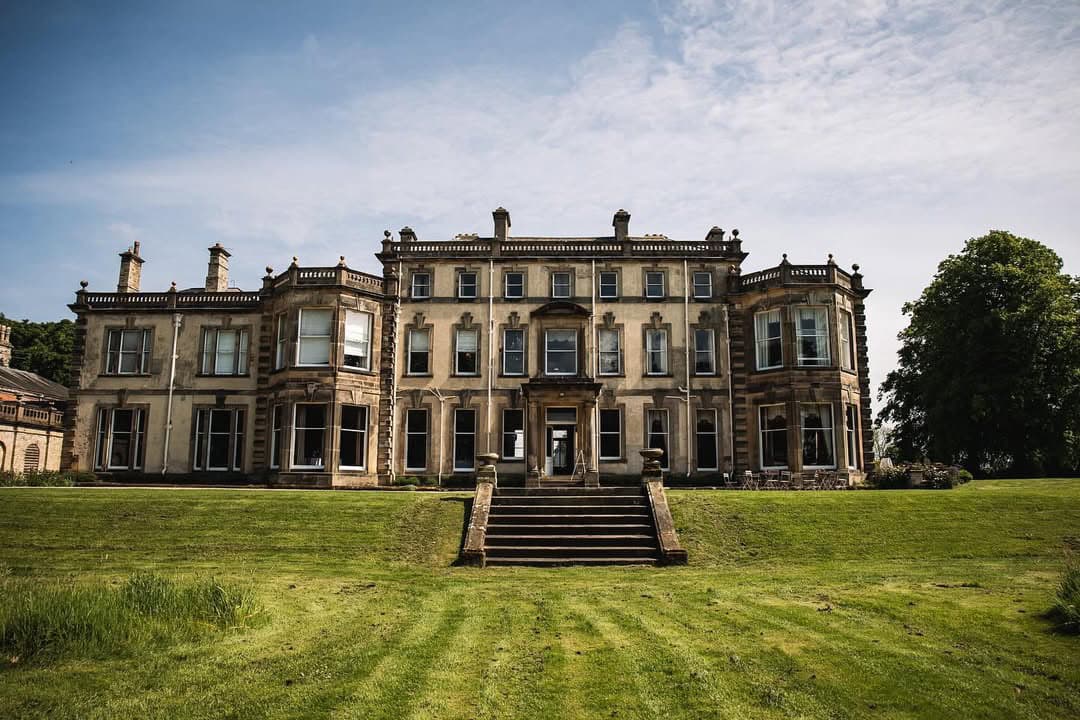 Grand stone building with multiple windows, steps leading to the entrance, surrounded by a green lawn and trees.