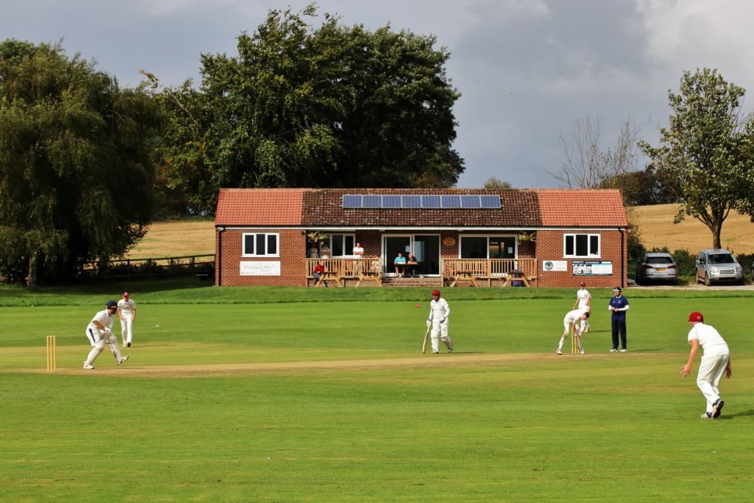 Cricket match in progress on a green field with a club house featuring solar panels in the background.