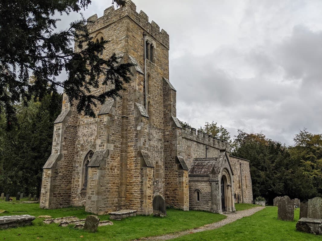 Historic stone church with a tall tower, surrounded by greenery and gravestones under a cloudy sky.