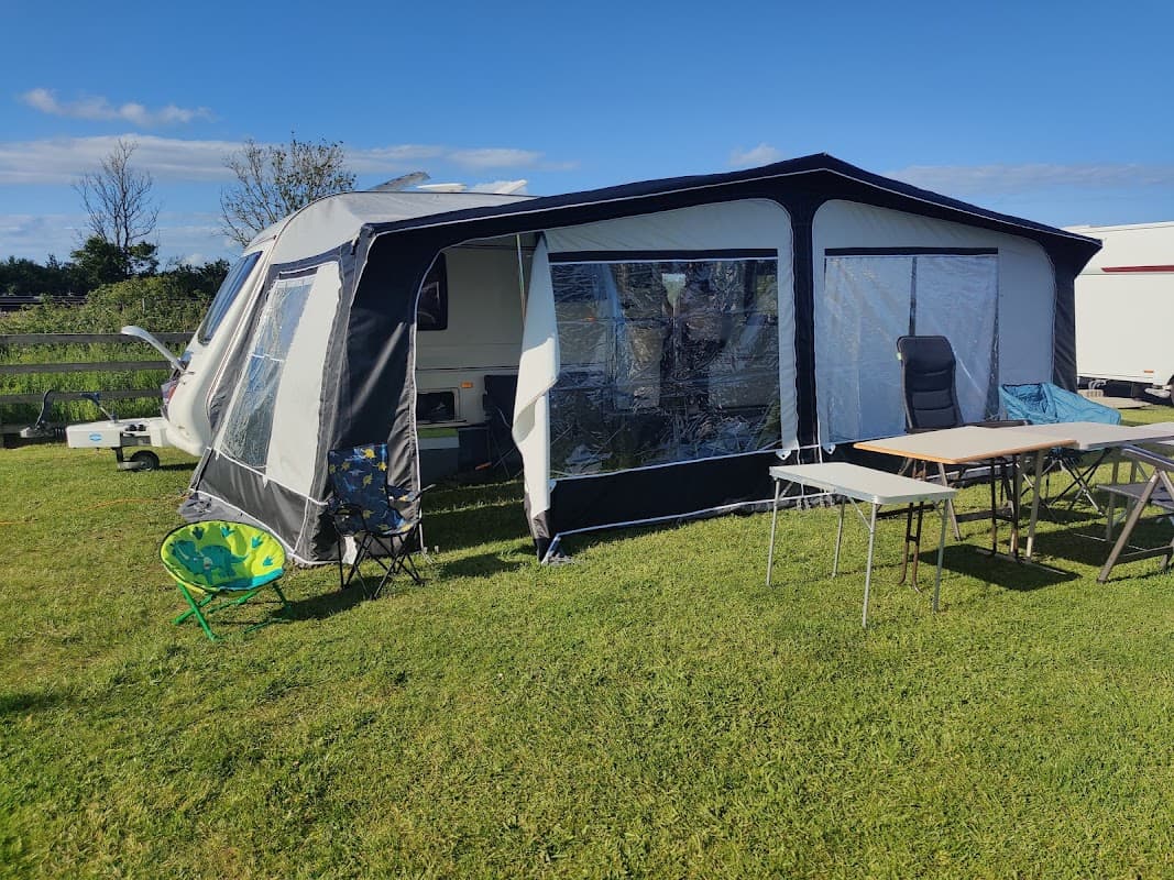 Caravan with an awning set on green grass, featuring outdoor chairs and tables under a clear blue sky.