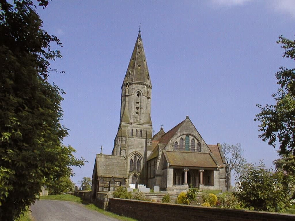 Historic stone church with a tall spire, surrounded by trees and a clear blue sky in East Heslerton, Yorkshire.