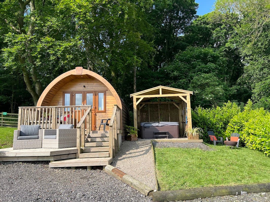 Wooden glamping pod with steps, surrounded by greenery, and a gazebo with a hot tub nearby on a gravel path.