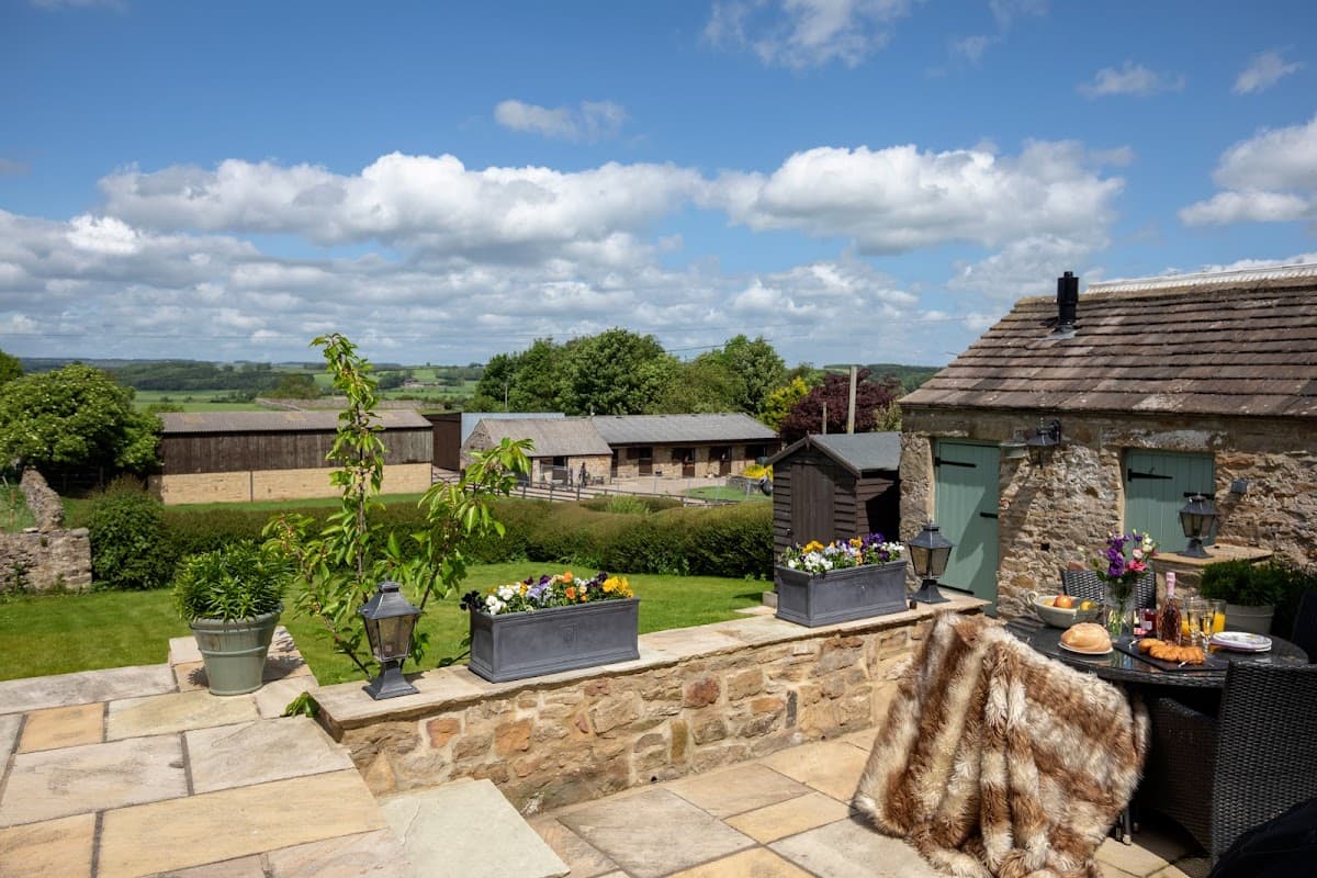 Scenic view of Manor House Cottages with stone walls, lush greenery, and flower planters under a bright blue sky.