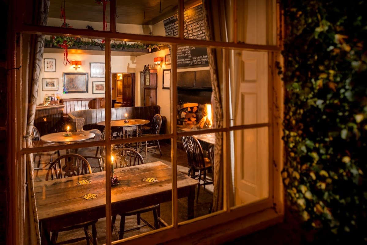 Cozy pub interior with wooden tables, chairs, a fireplace, and warm lighting, seen through a window.