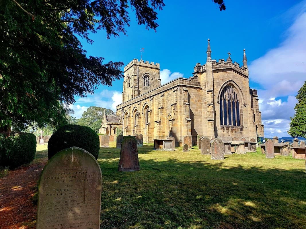 Historic stone church with tall tower, surrounded by gravestones and lush greenery under a bright blue sky.
