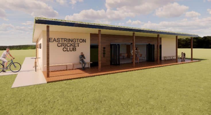 Modern cricket club building with a sign, outdoor seating, and people enjoying the space on a grassy field.