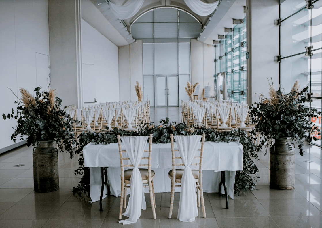 Elegant wedding setup with white drapes, greenery, and rustic chairs arranged in a spacious, modern venue.