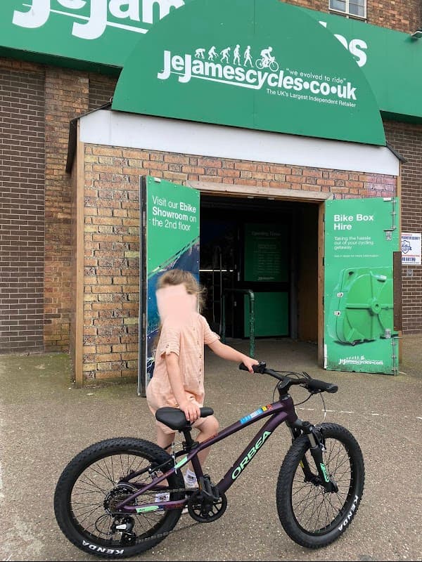 Child with a bike stands outside J E James Cycles shop, featuring a green sign and a bike hire display.