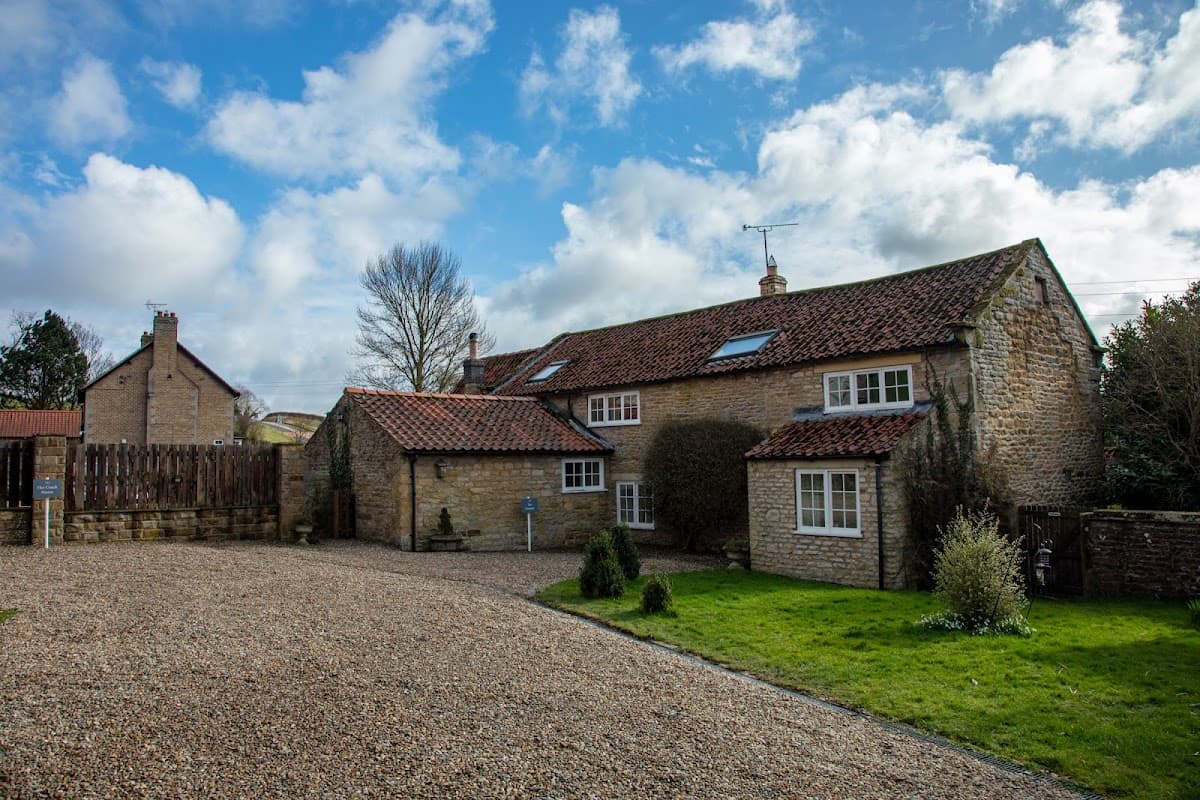 Stone cottages with a red-tiled roof, surrounded by green grass and gravel, under a partly cloudy sky.