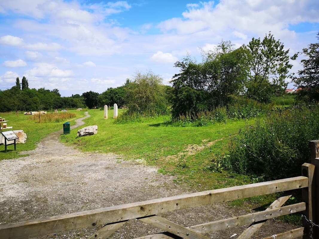 Lush green landscape with a gravel path, stone markers, and wooden gate under a blue sky with fluffy clouds.