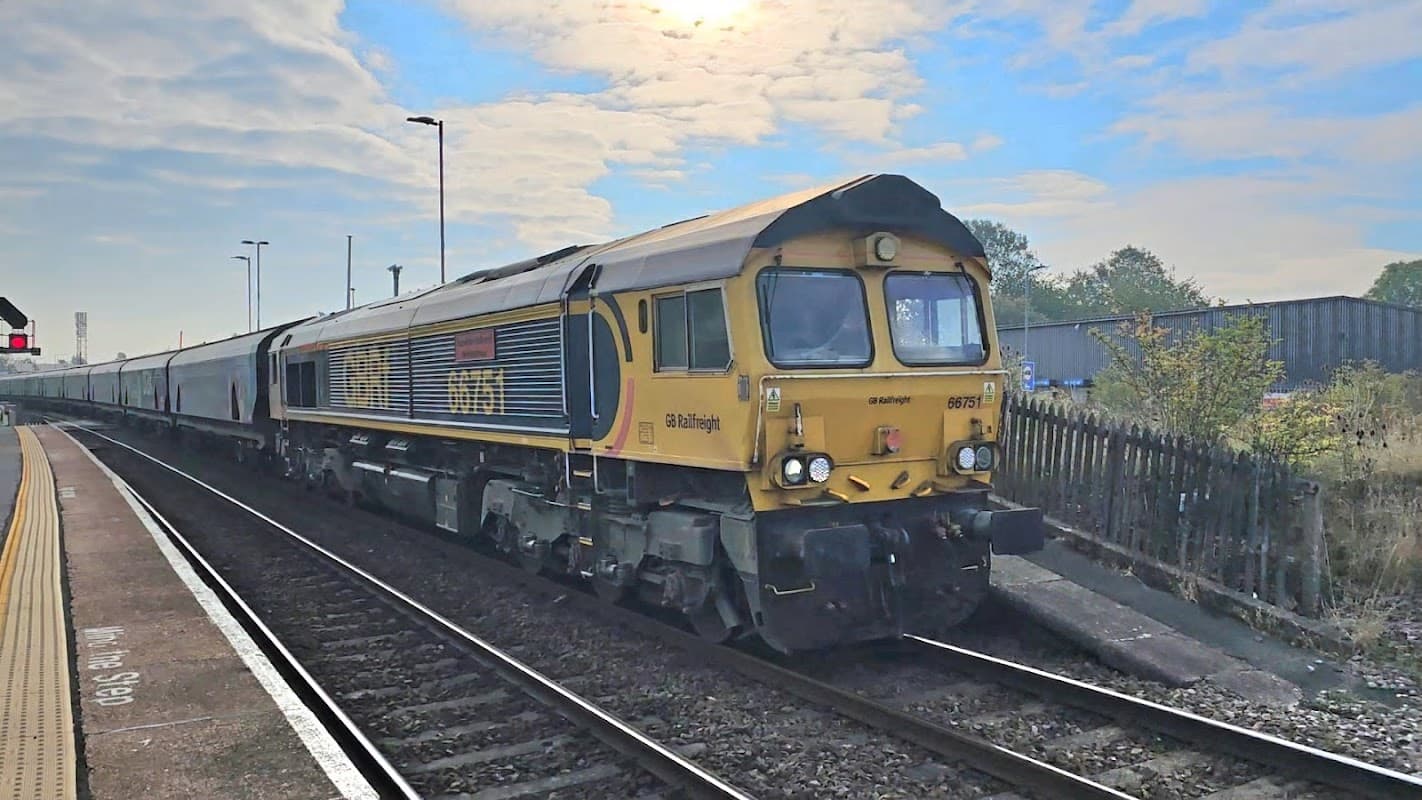 A freight train with a yellow and blue locomotive at Whitley Bridge Station, surrounded by tracks and cloudy sky.