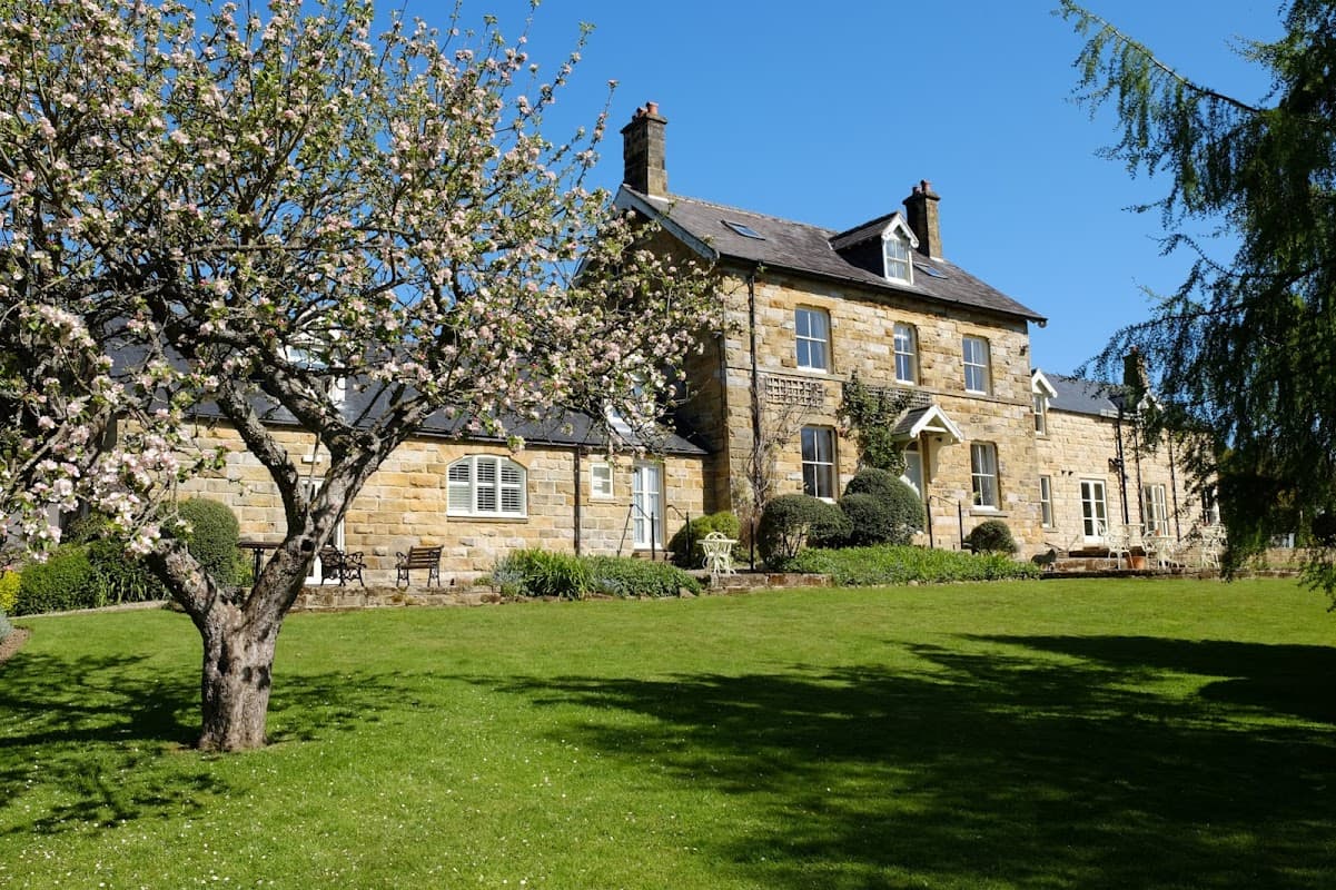 Broom House features a stone exterior, lush green lawn, flowering tree, and bright blue sky in Egton Bridge, Yorkshire.