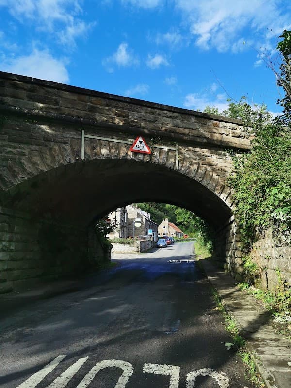 Stone bridge arching over a narrow road, with a warning sign and quaint buildings in the background under a blue sky.