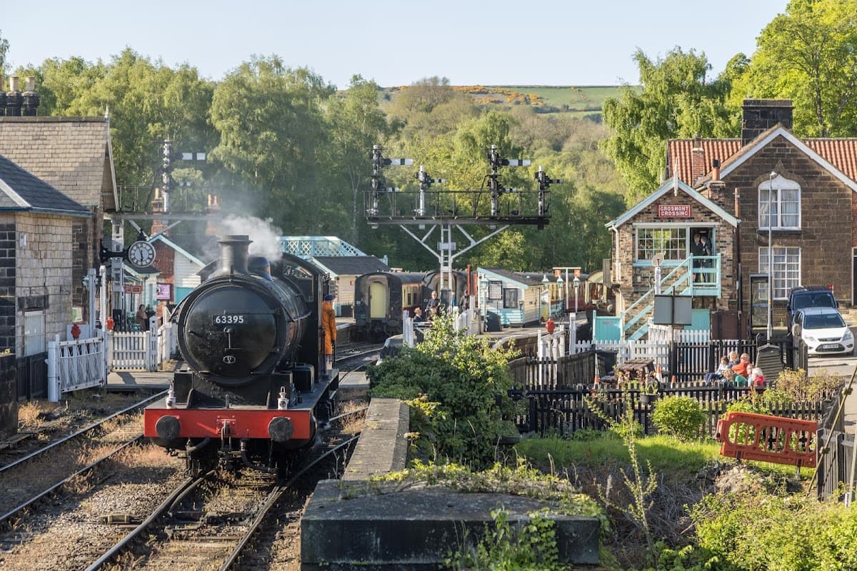 Steam train arriving at Grosmont station, surrounded by lush greenery and quaint buildings, with people enjoying the scenery.