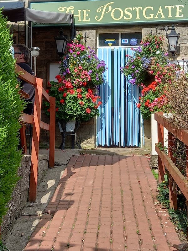 Colorful flower baskets adorn the entrance of Postgate Inn, with a brick path leading to the blue-striped door.