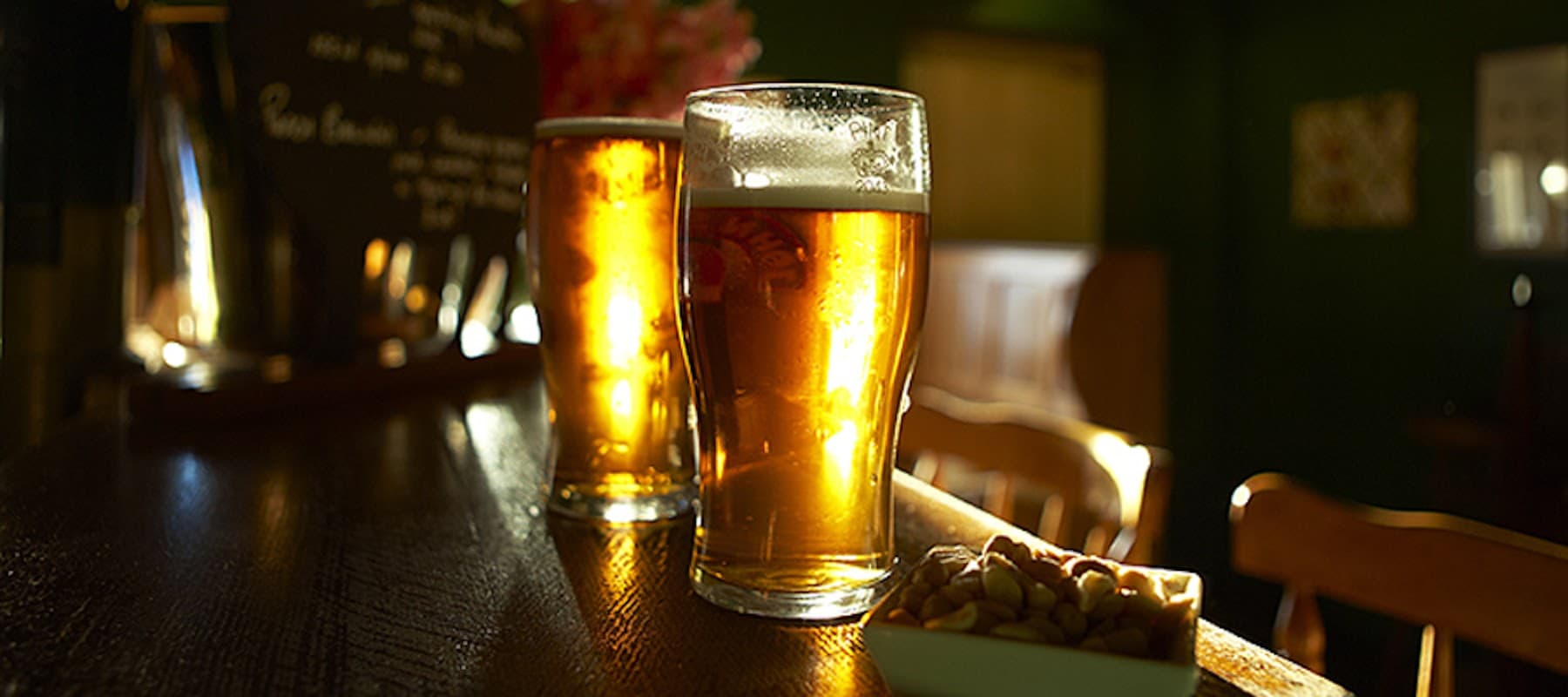 Two pints of golden beer on a wooden bar, with a bowl of snacks and a dark green pub interior in the background.