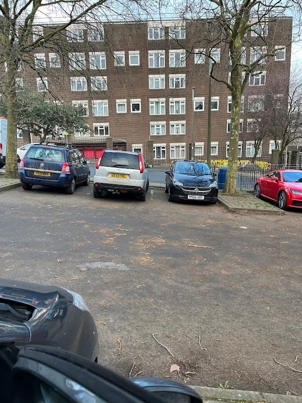 Free car park with several parked cars and a multi-story building in the background, surrounded by bare trees.