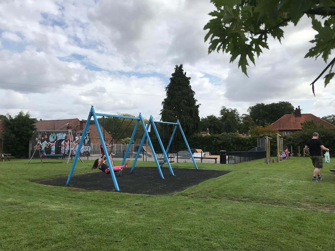 Colorful swings in a grassy park, with a child swinging and a man nearby, surrounded by trees and buildings.