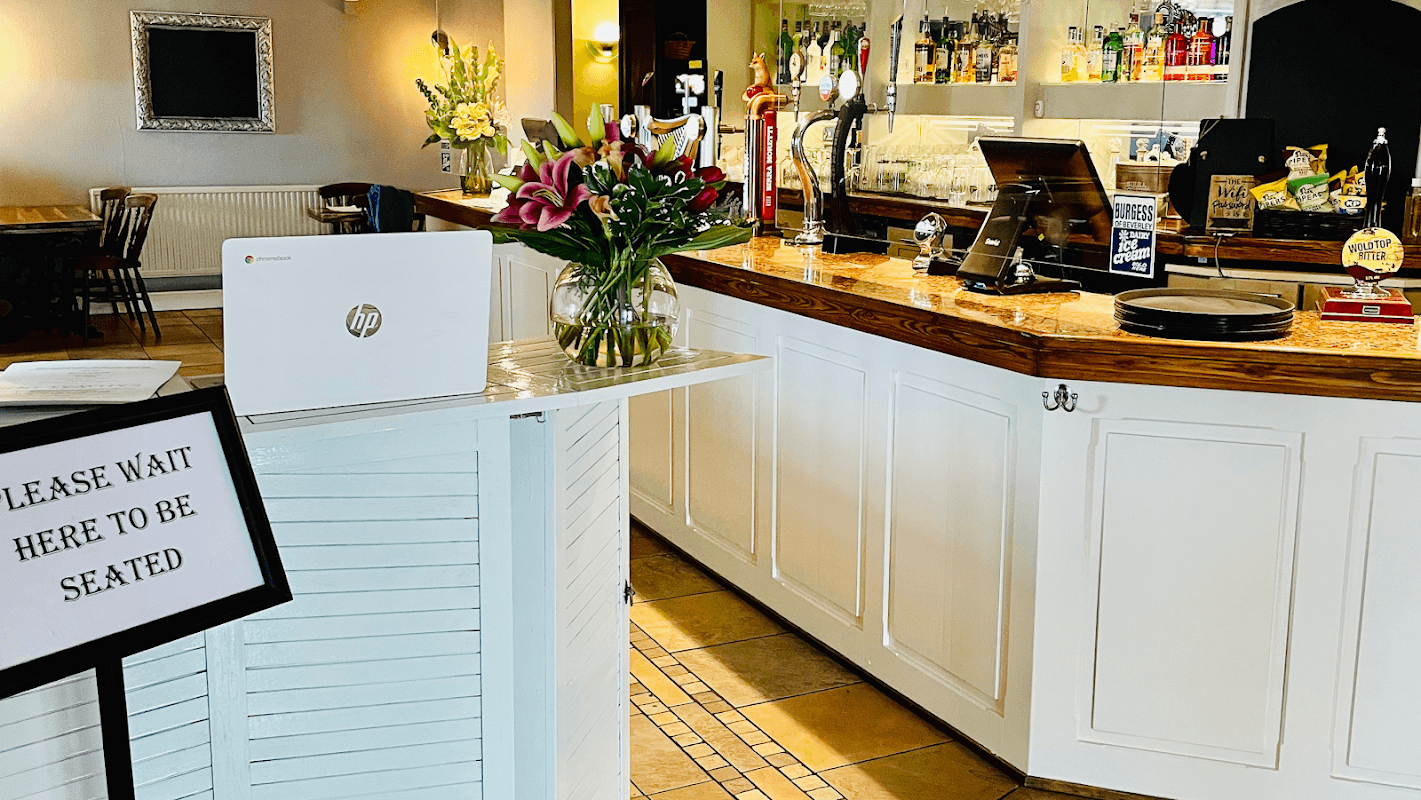 Cozy pub interior with a bar, colorful drinks, fresh flowers, and a sign requesting patrons to wait to be seated.