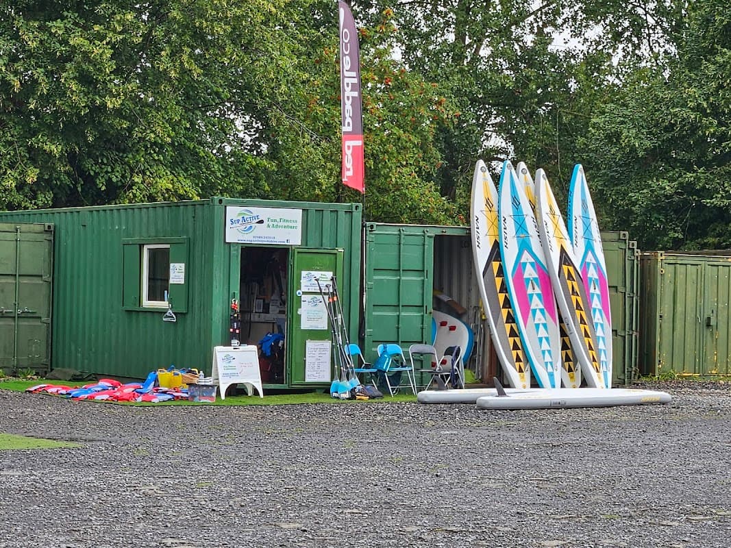 Green shipping container with paddleboards outside, surrounded by trees and equipment for water sports.