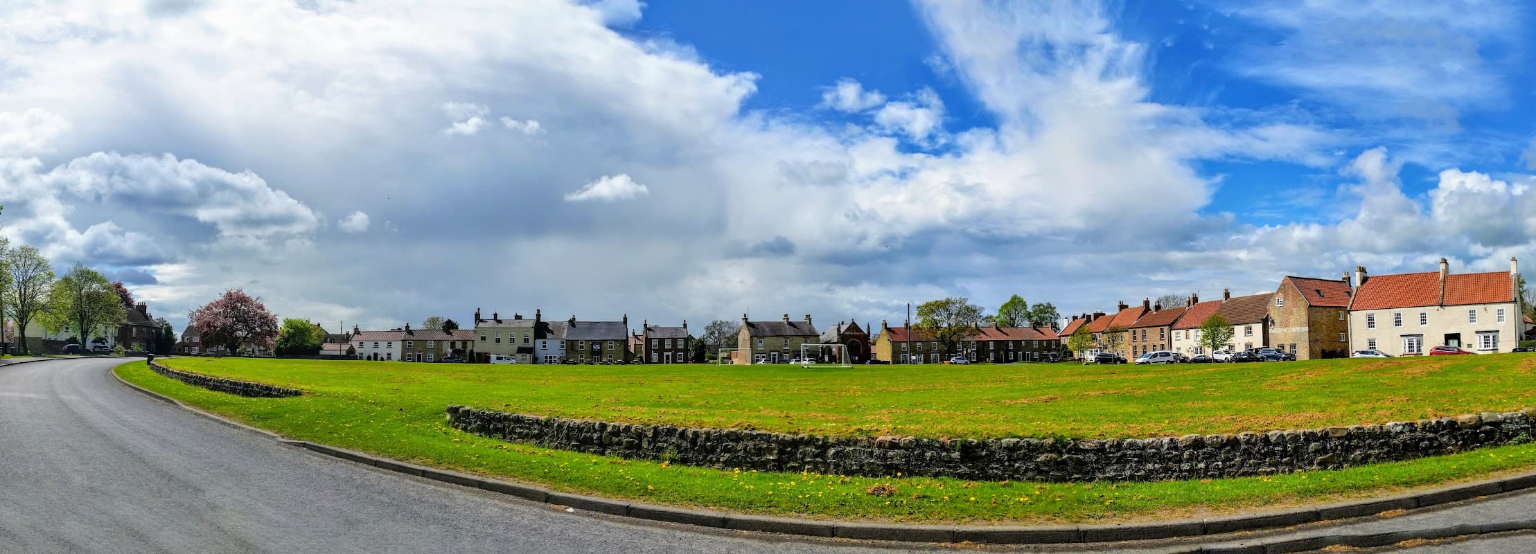 A panoramic view of The Green in Ellerton-on-Swale, featuring cottages, a grassy area, and a blue sky with clouds.