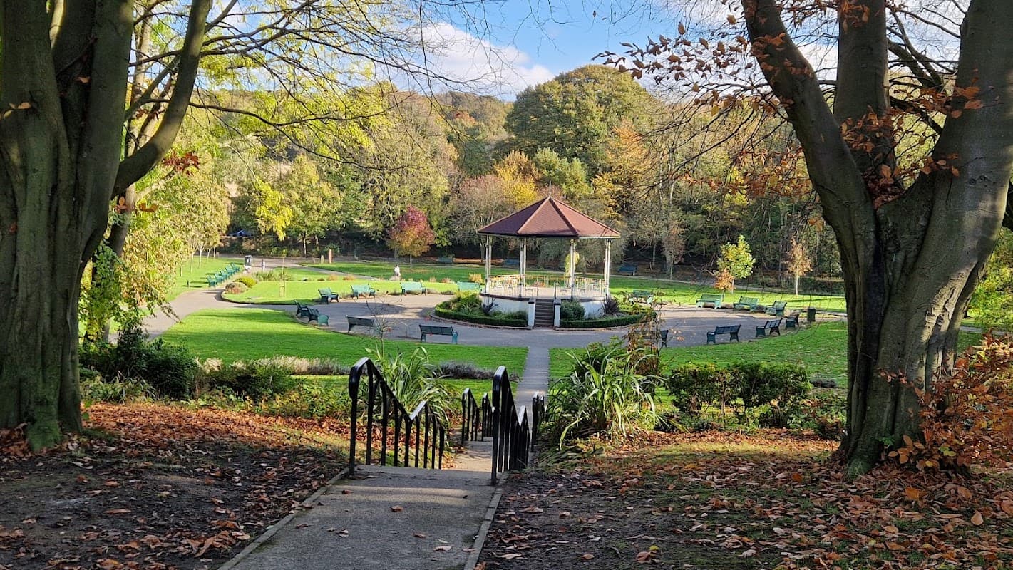 A path leads down to a circular garden with a gazebo, surrounded by trees and benches in autumn colors.