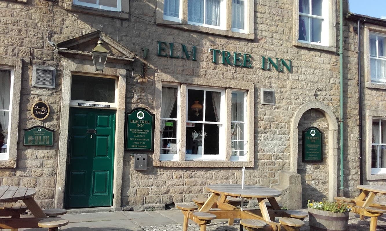 Stone facade of Elm Tree Inn with green door, windows, and wooden benches outside in Embsay, Yorkshire.