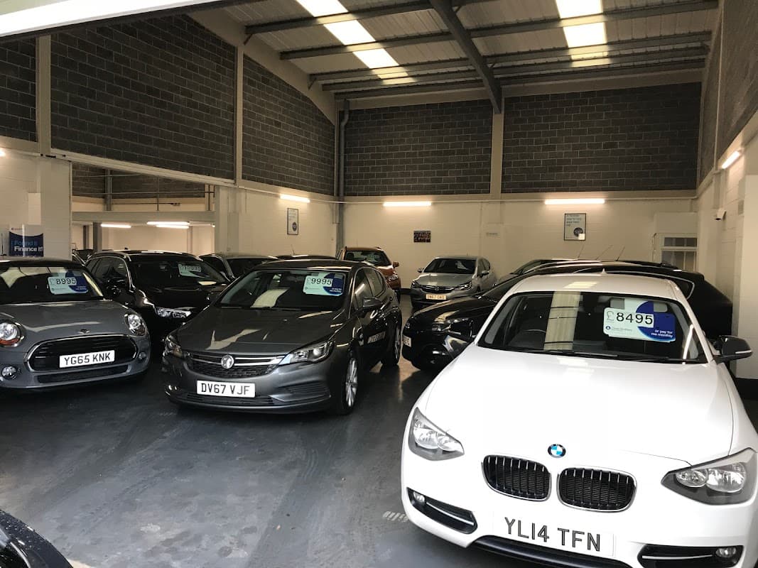 Car dealership interior with various models on display, price tags visible on several vehicles.