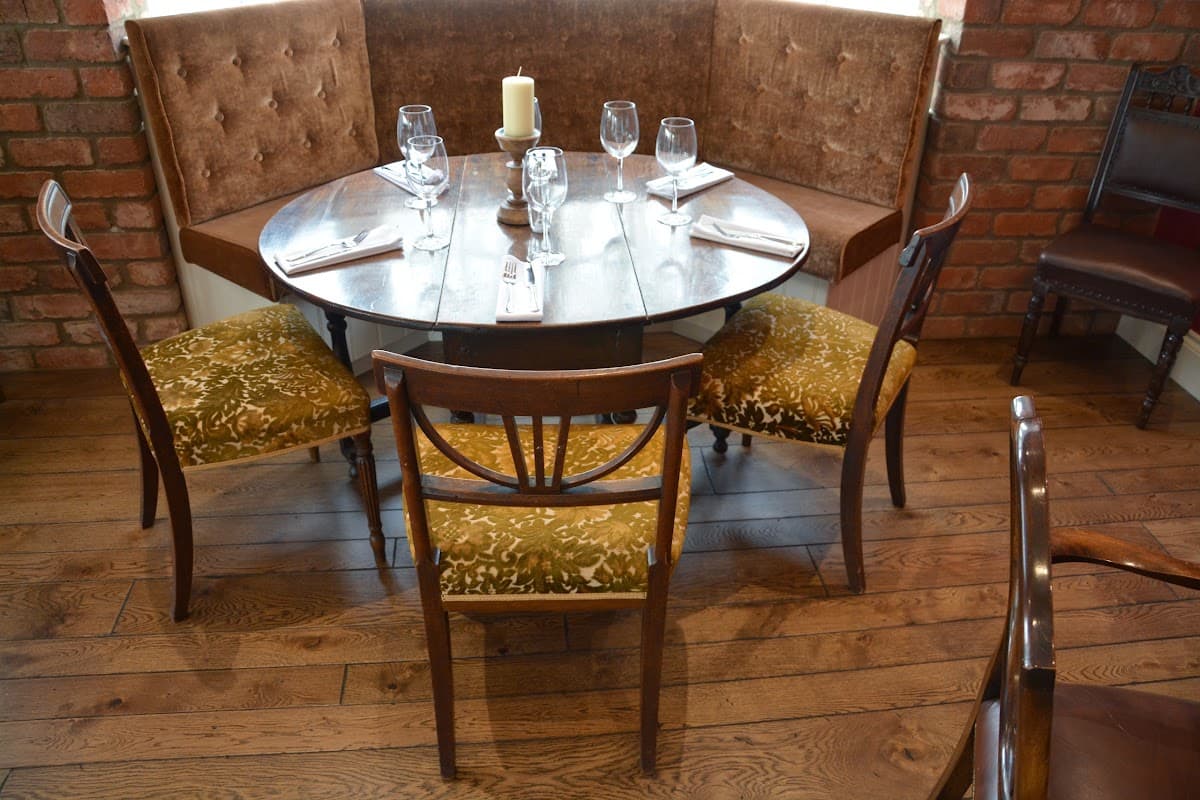 Cozy dining area with a round wooden table, elegant glassware, and upholstered chairs against a brick wall.