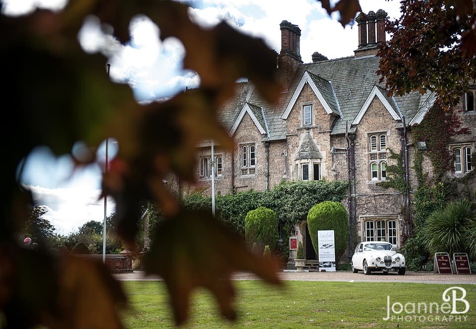 Historic building with ivy-covered walls, manicured lawns, and a vintage car in front, set against a cloudy sky.