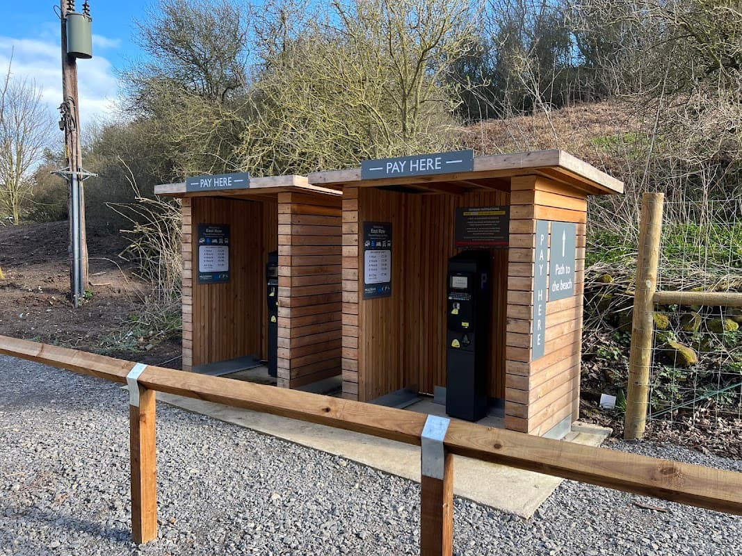 Two wooden pay stations with signage, surrounded by trees and gravel path in East Row Car Park, Whitby, Yorkshire.