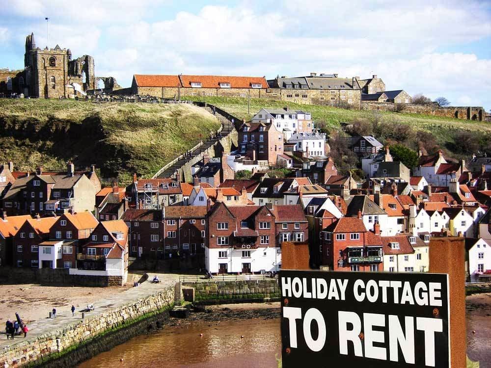 Holiday cottages with red-tiled roofs, a hilltop church, and a sign reading "Holiday Cottage To Rent" in Whitby.