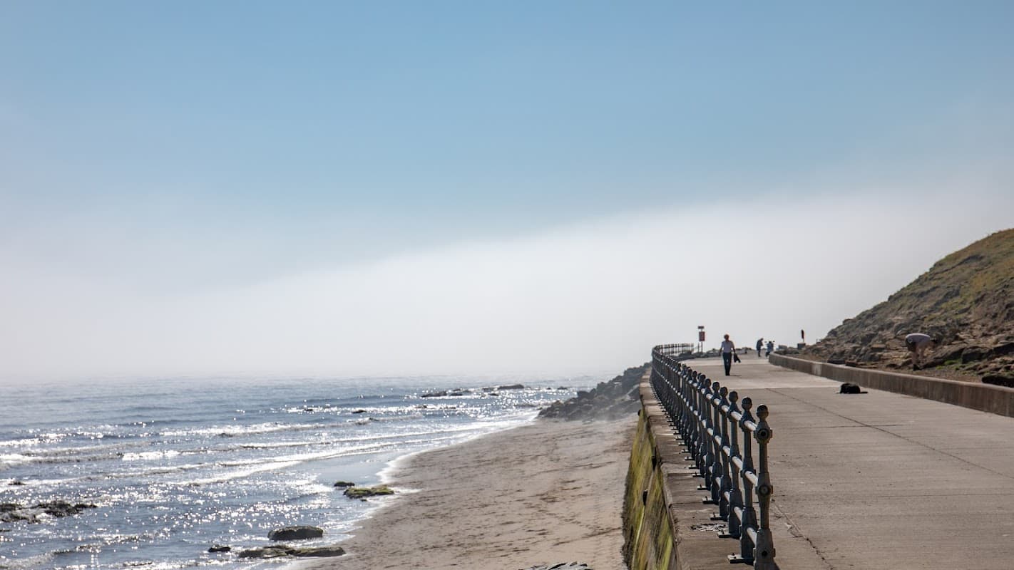 Coastal path alongside a sandy beach with gentle waves, under a clear blue sky and distant fog.