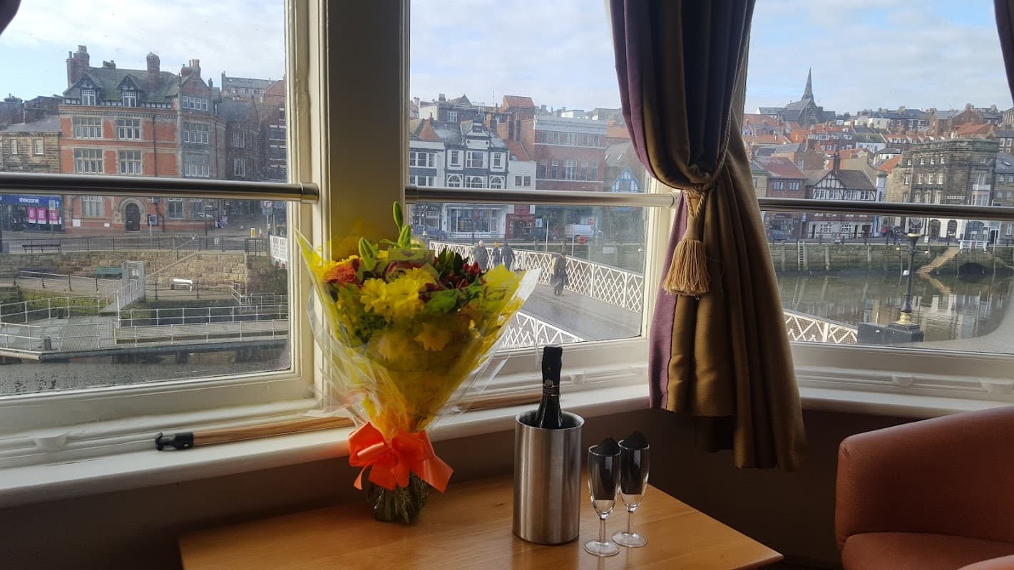 Bouquet of flowers and champagne glasses on a table by a window, overlooking Whitbyβs colorful buildings and harbor.