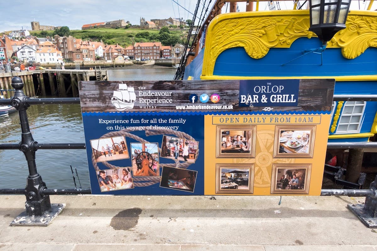 Colorful sign for "The Whitby Endeavour" bar with images and text, overlooking the harbor and buildings of Whitby.