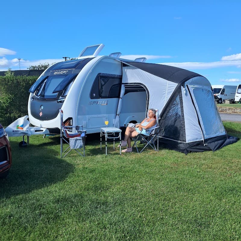 A man relaxes outside a caravan with a sunshade on green grass under a clear blue sky at Melgoats CL Site.