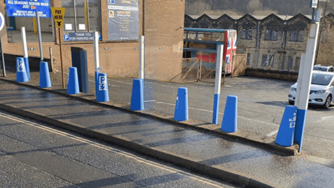Blue and white parking posts line a car park with a payment sign, near a building and a bus in the background.