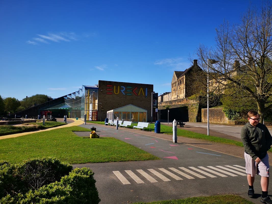 Pay & Display car park with colorful building, benches, and green space under a clear blue sky in Exley, Yorkshire.