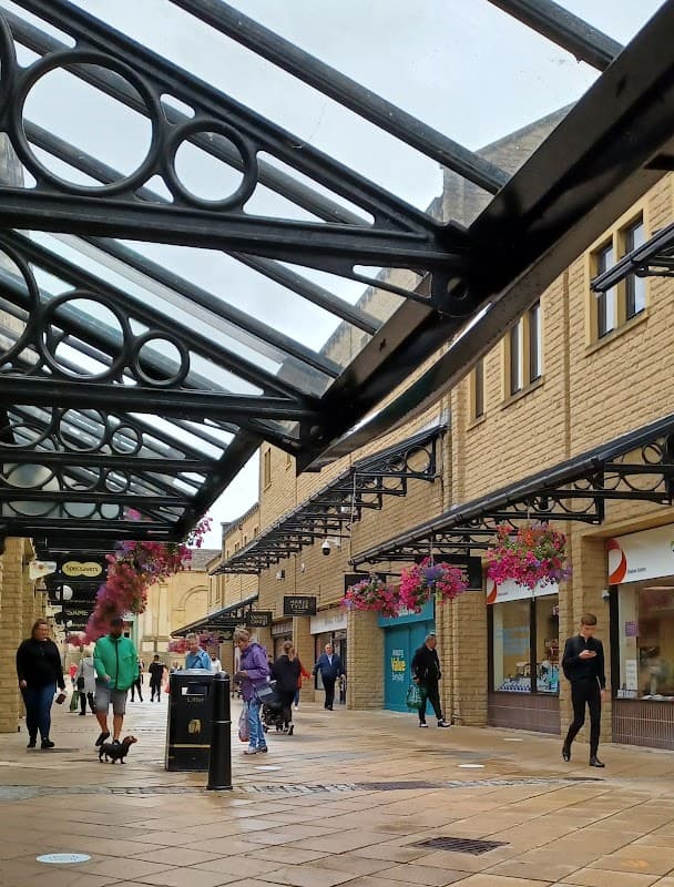 Covered walkway with shops, hanging flower baskets, and people walking in Woolshops Shopping Centre Car Park, Exley.