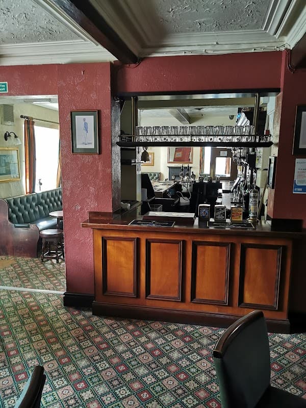 Wooden bar with glassware, green leather seating, patterned carpet, and warm lighting in a cozy pub interior.
