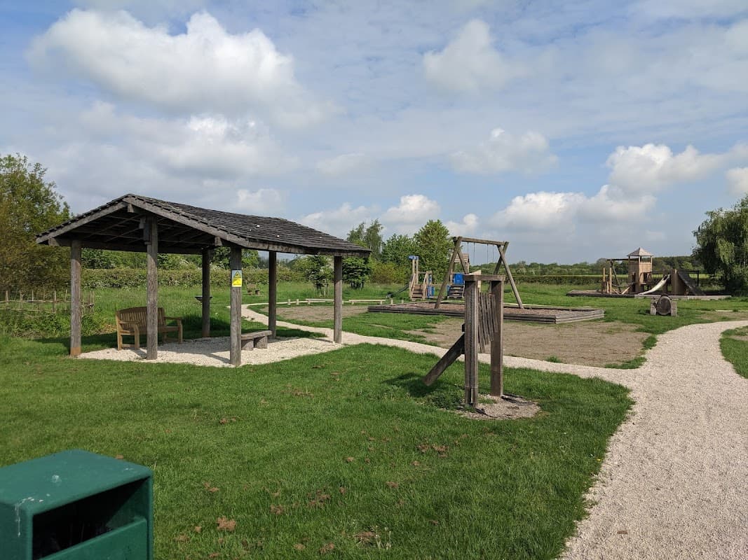 Wooden shelter with benches, swings, and play structures under a blue sky with fluffy clouds in Jubilee Park, Fangfoss.