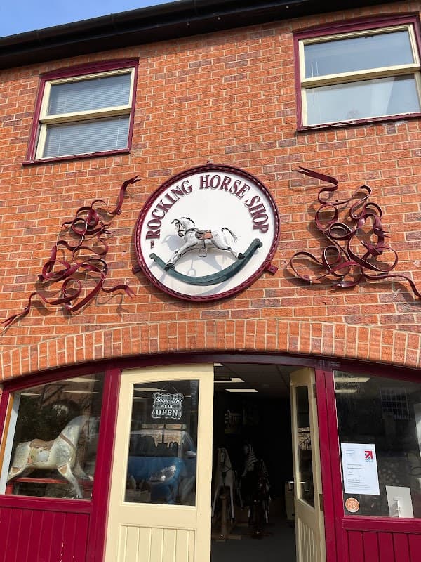 Sign for "The Rocking Horse Shop" on a brick building, featuring a rocking horse design and decorative swirls.
