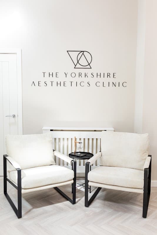 Two stylish white armchairs with a small black table between them, featuring the Yorkshire Aesthetics Clinic logo on the wall.